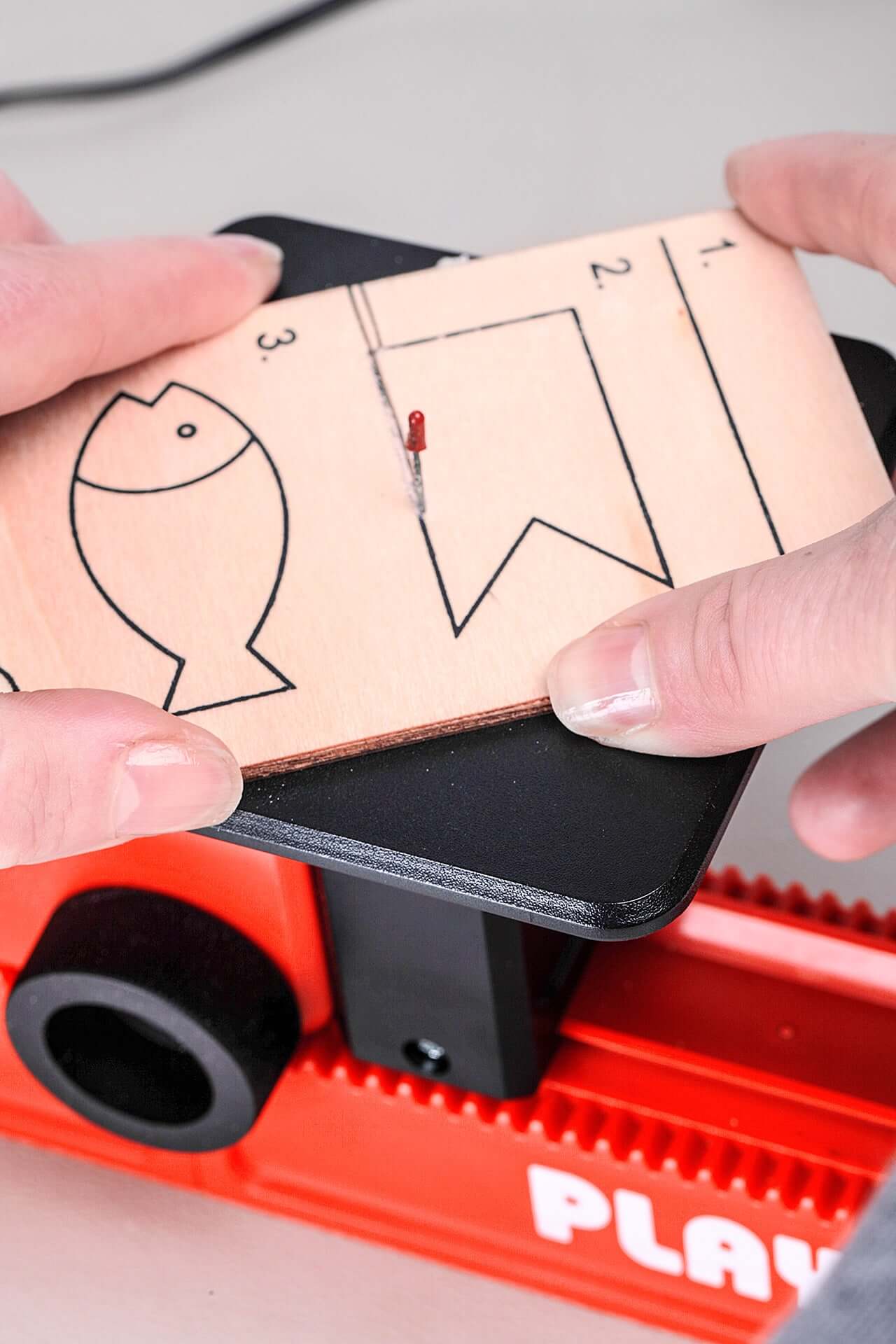 Hände führen eine Holzplatte mit aufgezeichneter Kontur an der PLAYmake-Säge.